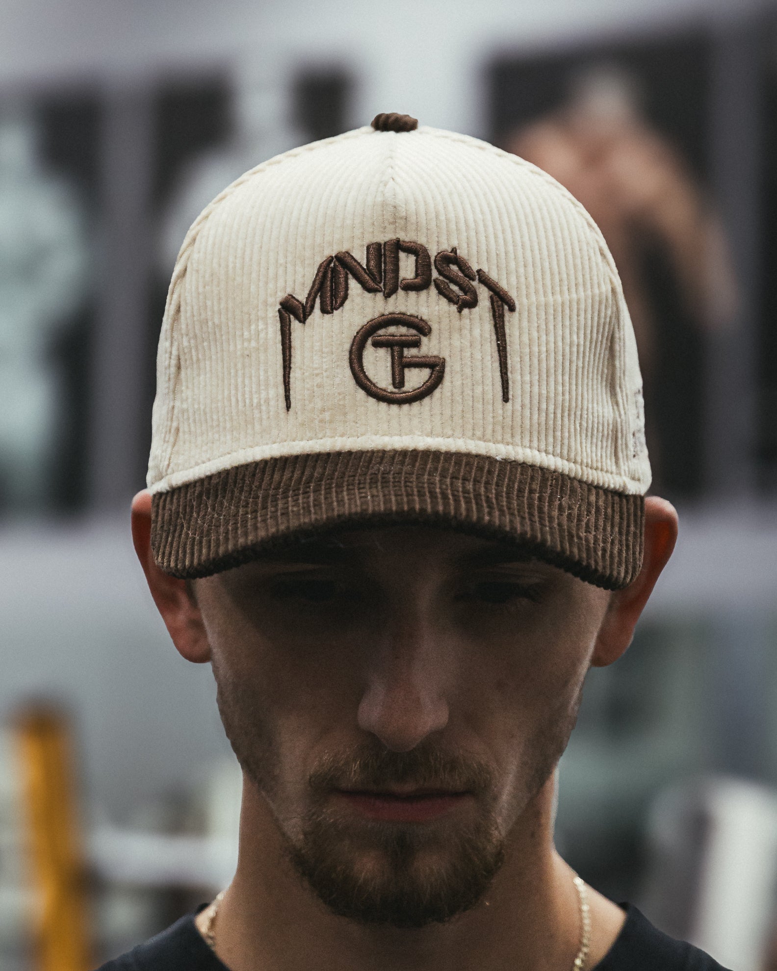 Man wearing a cream and brown corduroy MNDST TG Collab cap with text on a blurred background
