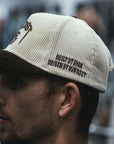 Man wearing a cream and brown corduroy MNDST TG Collab cap with text on a blurred background