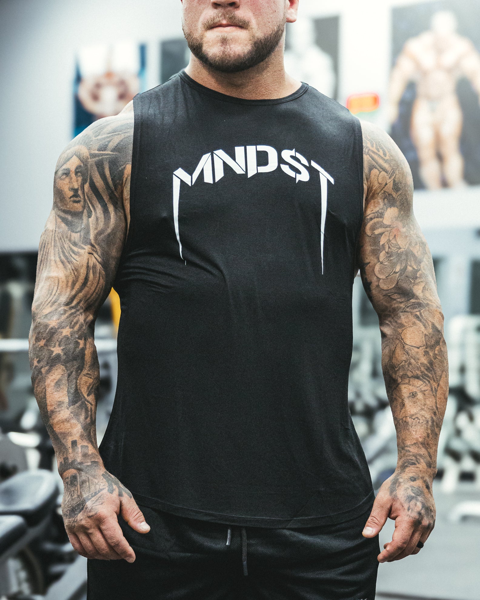 Man wearing a black sleeveless shirt or tank top with 'MNDST' logo in a gym setting