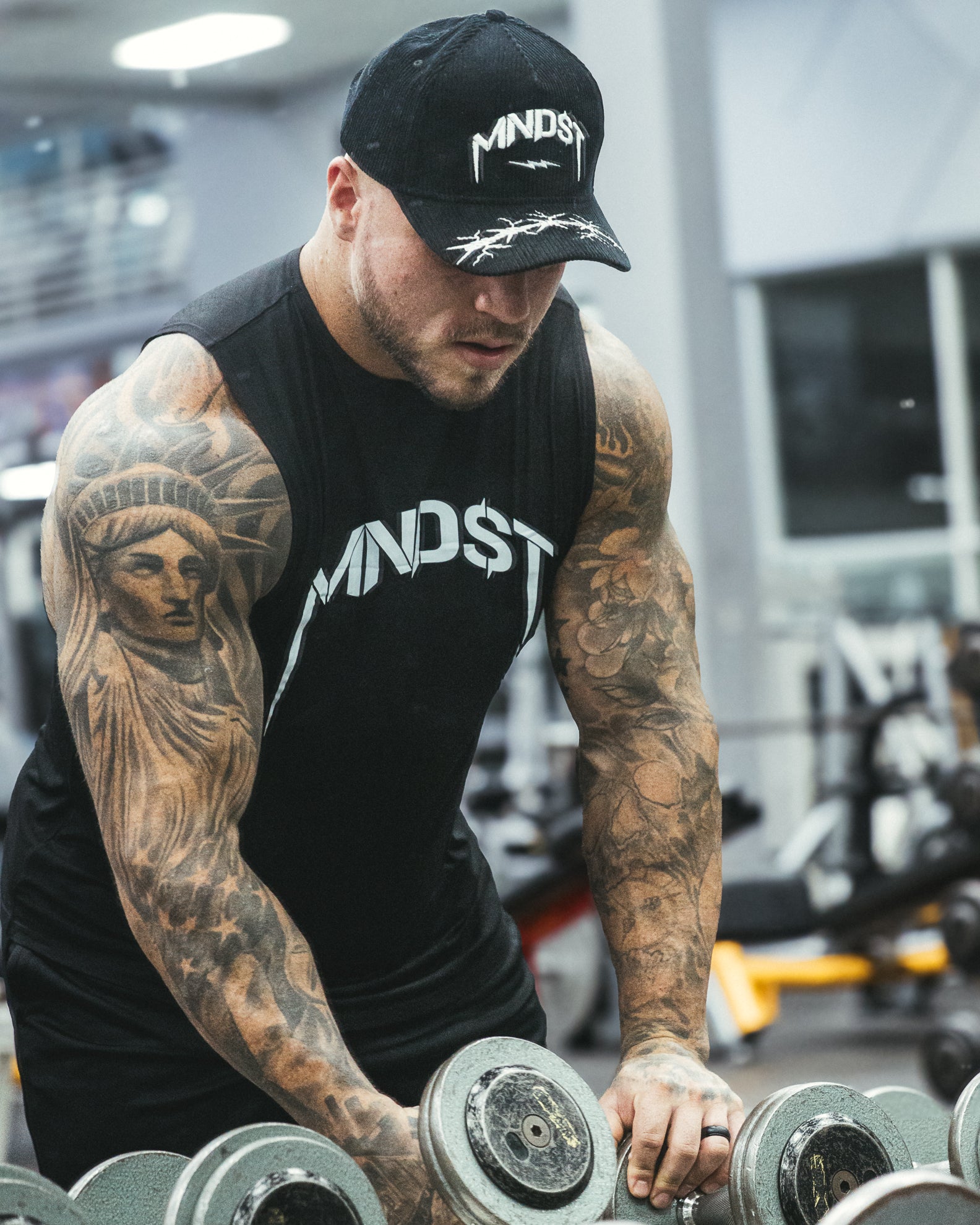 Man lifting weights in a gym wearing a black 'MINDST' cap and tank top.