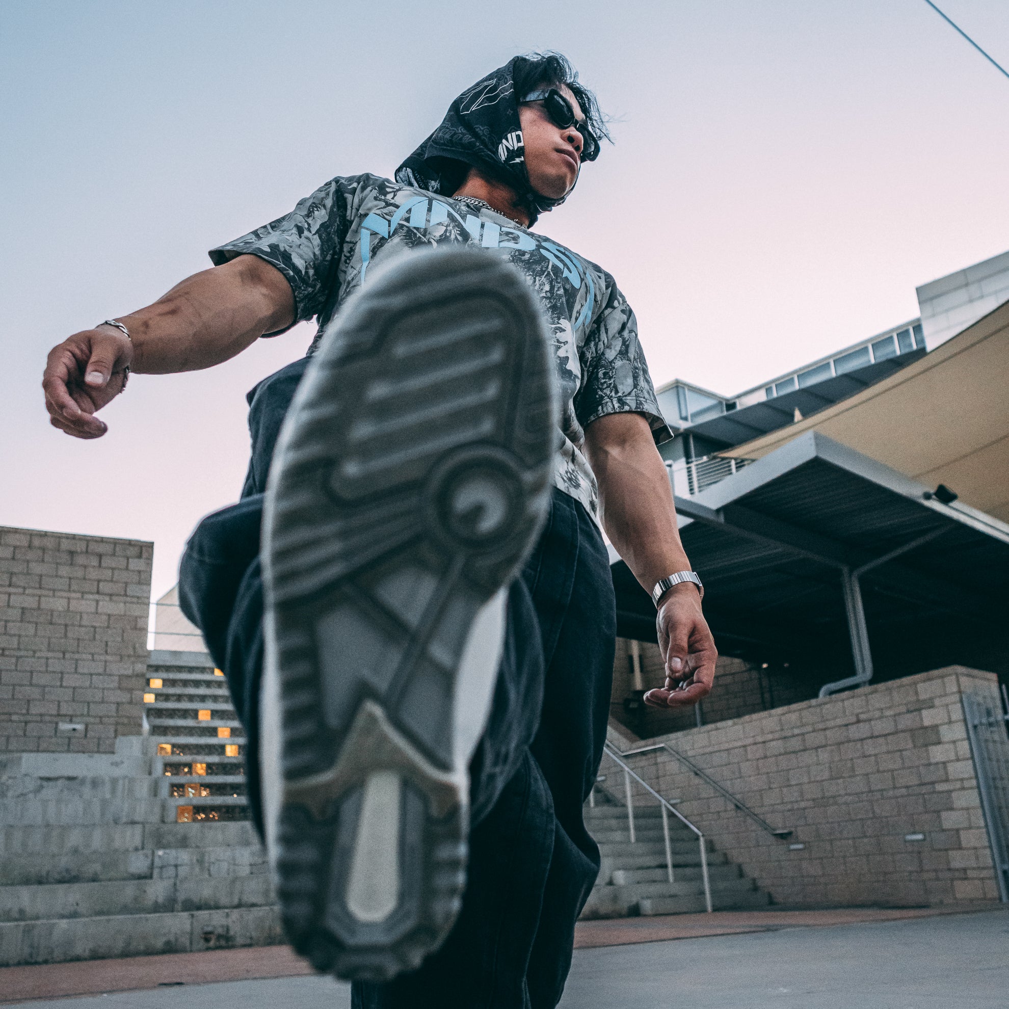 Person wearing a MNDST camouflage shirt and black pants with a focus on their shoes in an urban setting.