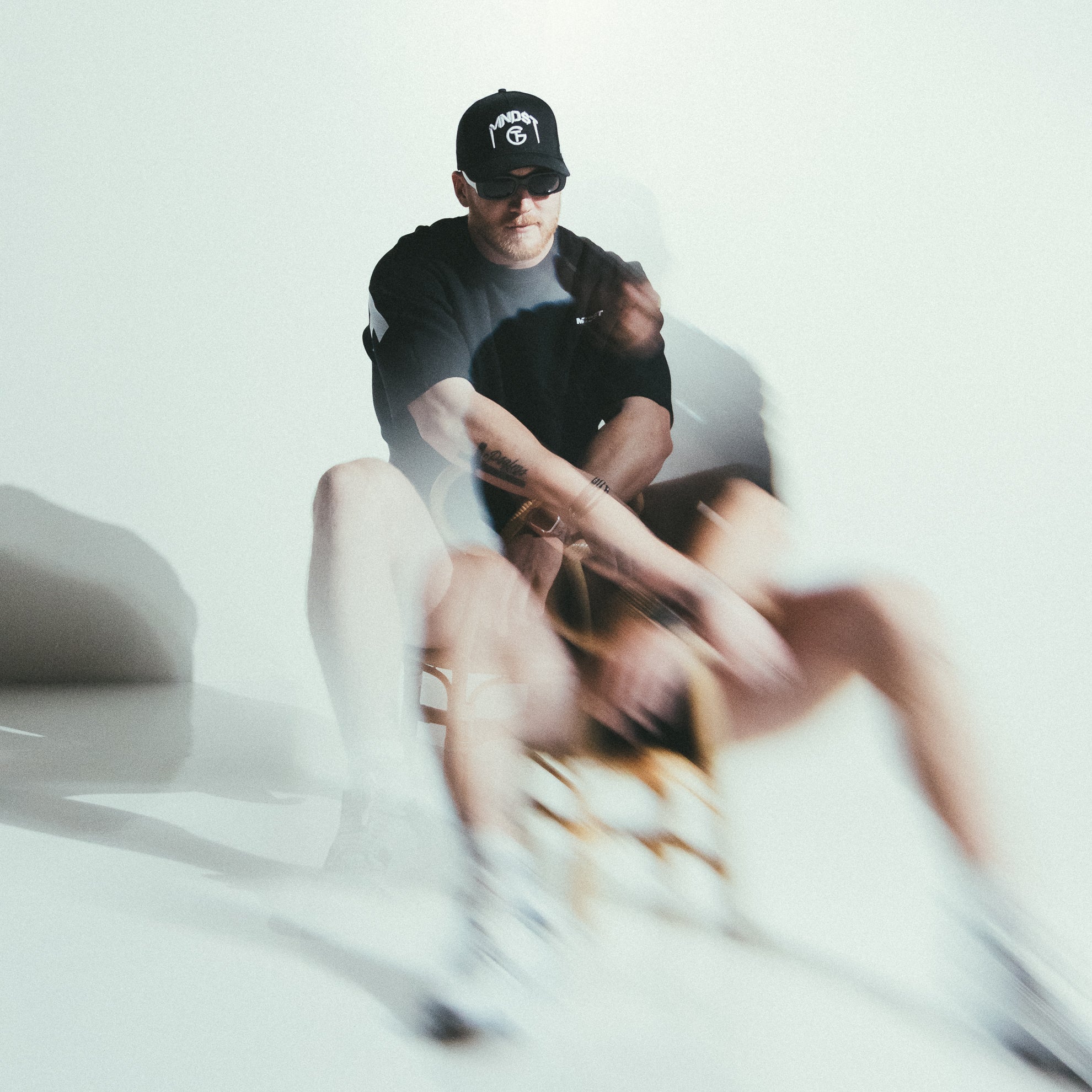 Person wearing a black MNDST cap and sunglasses sitting on a white surface with a blurred background